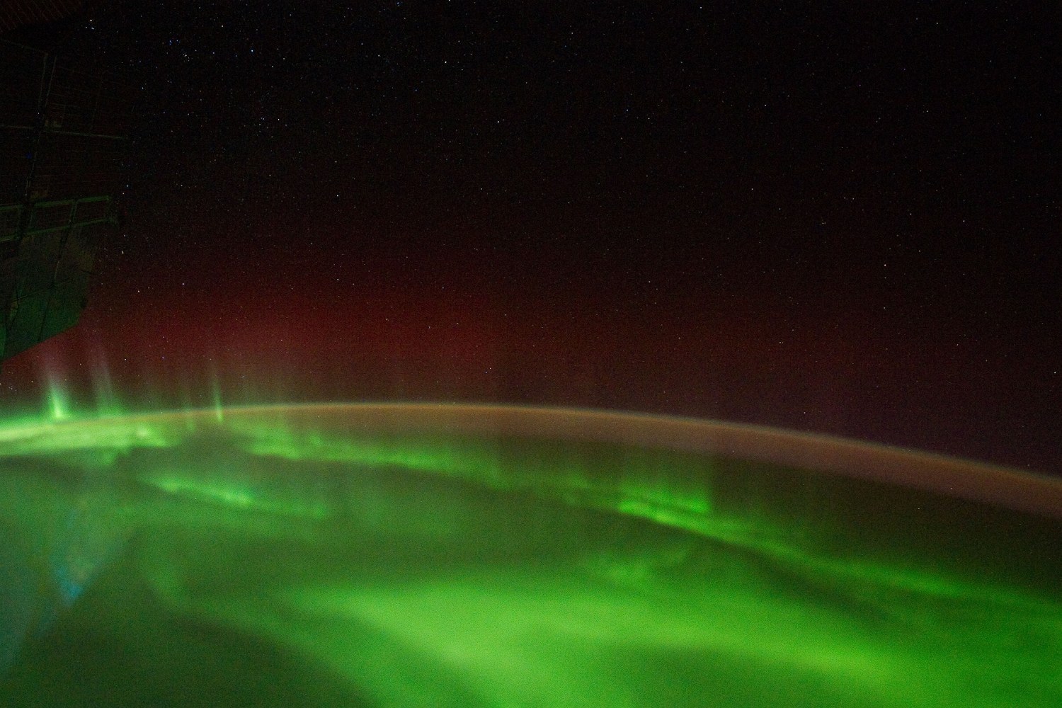 Curtains of green aurora with red fringes over a planet's night side, seen from orbit.