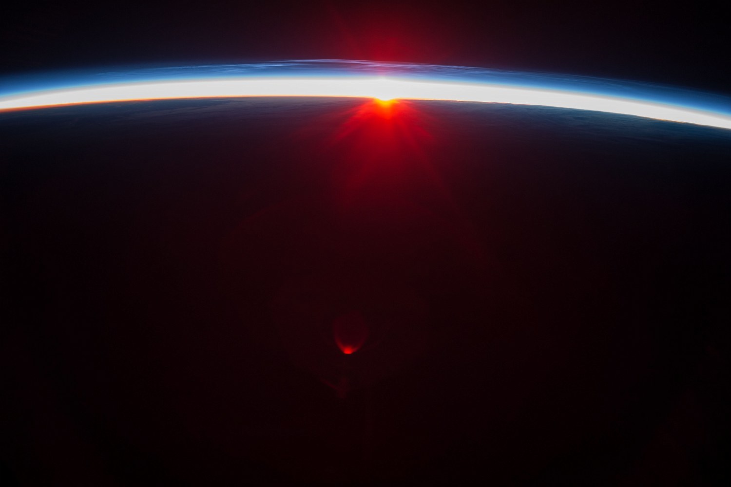 A red sunrise breaking over a planet's thin, bright blue band of atmosphere, seen from orbit.