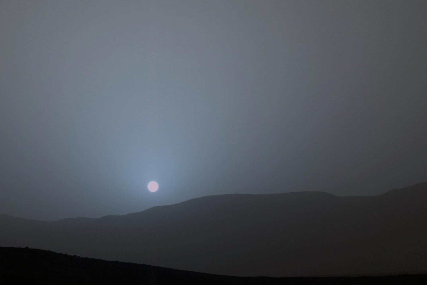 A small pale sun setting in a blue glow over a dark ridge, the sky grey-brown.