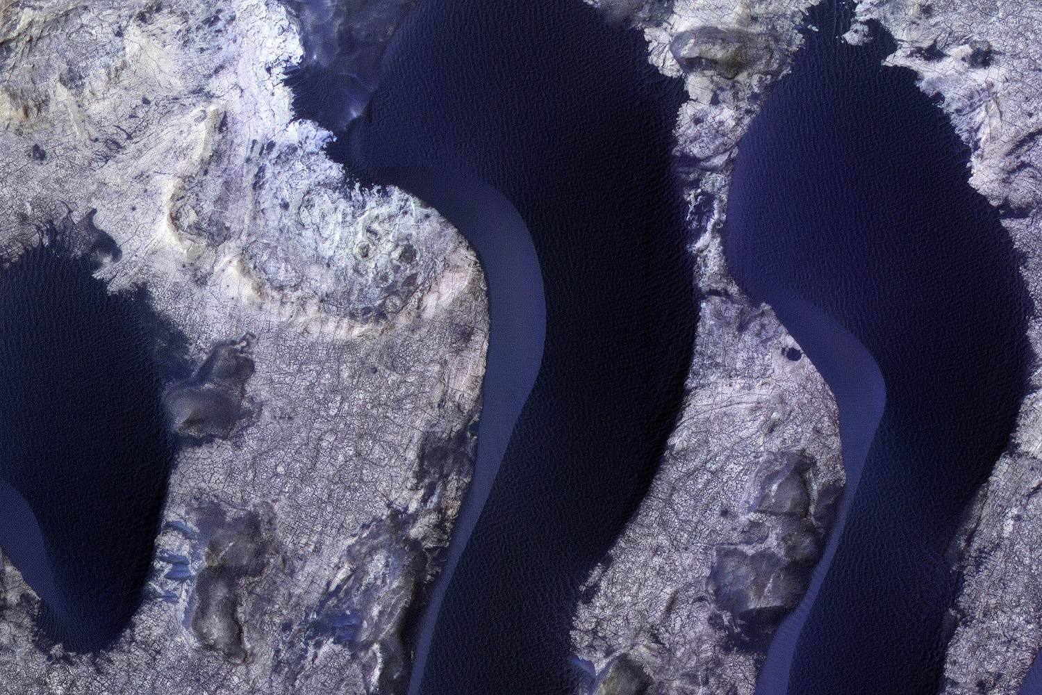 Seen from above, dark blue-black dunes curving in long arcs across pale, cracked ground.