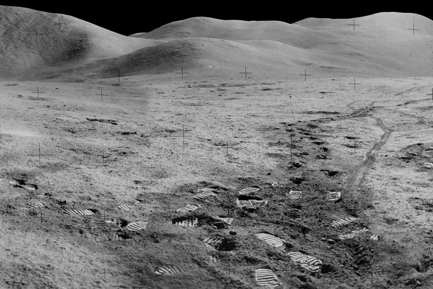 Grey rolling lunar hills under a black sky, boot prints and tracks in the dust in the foreground.